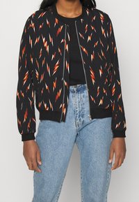 Černá bombarďácká bunda se vzorem blesku v oranžové a bílé barvě. Obsahuje žebrované manžety a přední zip. Nositá přes černý top.