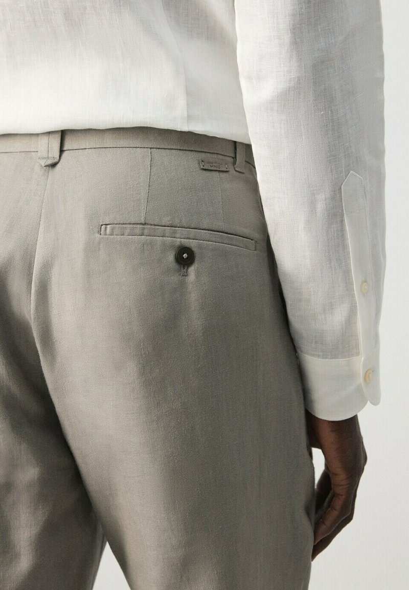 Gros plan sur une personne portant un pantalon beige ajusté avec une poche arrière boutonnée et une chemise blanche à manches longues avec des boutons aux poignets.