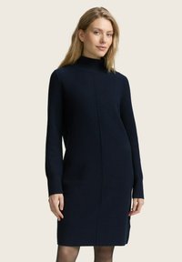 Gebreide donkerblauwe jurk met een funnelhals, lange mouwen en een rechte silhoeet. Heeft een centrale naaddetail en een geribbelde textuur.