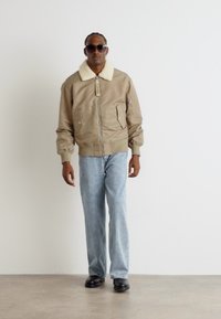 Chaqueta bomber beige con cuello de pelaje sintético, cierre frontal y dos bolsillos laterales, combinada con jeans de color azul claro y zapatos negros.