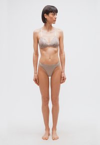 Γυναίκα με κοντά σκούρα μαλλιά, όρθια ξυπόλυτη, φορούσα μπεζ δαντελένιο σουτιέν και εσώρουχα που ταιριάζουν, κοιτάζοντας προς τα αριστερά της πάνω σε απλό φόντο.