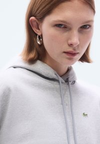 Helehall hall kapuutsiga dressipluus paelakestega, millel on rinnal väike roheline logo. Sile tekstuur, kantud hõbedaste rõngakõrvarõngastega.