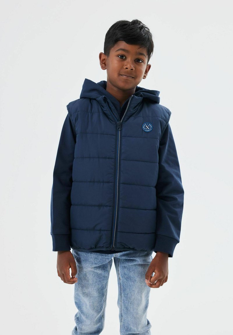 Marinblå vadderad väst med huva, med en framdragkedja och en cirkulär logotyp. Bärs över en mörk topp, i kombination med ljusa denimjeans.