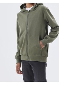 Groene ritshoodie met een kangoeroe-pocket, ribgebreide manchetten en zoom. Zachte stof met een capuchon, gedragen over een wit t-shirt, gecombineerd met donkere broek.