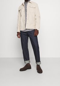 Beżowa bawełniana kurtka koszulowa, szara koszula zapinana na guziki, ciemne spodnie dżinsowe podwinięte przy mankietach, brązowe skórzane buty, stojące na białym tle.