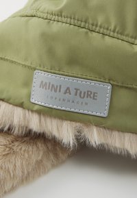 Tissu matelassé vert olive avec bordure en fausse fourrure beige, comportant une étiquette rectangulaire argentée embossée du nom de la marque "MINI A TURE COPENHAGEN."