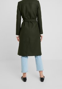 Cappotto verde oliva con cintura, colletto a scialle e maniche lunghe, silhouette dritta e schiena ventilata, abbinato a jeans azzurri chiari e scarpe nere.