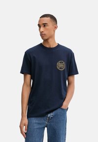 Camiseta de algodón azul marino con cuello redondo y mangas cortas. Presenta un logotipo circular dorado en el lado izquierdo del pecho. Combinada con jeans de mezclilla azul claro.