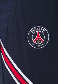 Granatowe krótkie spodenki sportowe z okrągłym logo Paris Saint-Germain oraz czerwono-białymi paskami po bokach. Gładka syntetyczna tkanina.