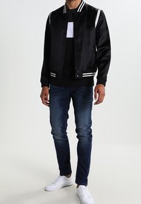 Svart satin bomberjacka med vita ränder på axlarna, bärs över en svart och vit skjorta, matchat med blå jeans och vita sneakers.