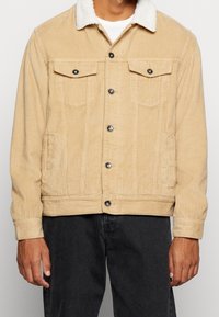 Beige manchesterjacka med vit fuskpälskrage, två bröstfickor framtill och svarta metalldetaljer, med räfflad textur.