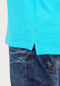 Ljust blå polo skjorta med strukturerad yta, med sidoflikar vid sömmen, bärs över mörka denimjeans med synliga söm detaljer.