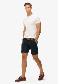 Navy shorts, hvid t-shirt, brun læderbælte og vinrøde bådsko. Outfitet har et simpelt, afslappet design med en tætsiddende pasform.