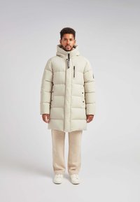 Giacca puffer beige chiaro con cappuccio, zip frontale, due tasche laterali e tasca sul petto con zip. Completata da pantaloni beige chiaro e sneakers.