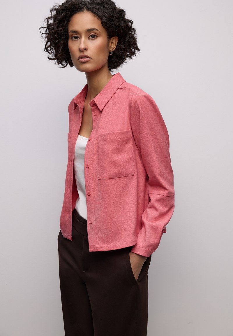 Chemise rose à boutons avec col et une poche poitrine, associée à un pantalon marron foncé. Texture lisse, coupe décontractée.