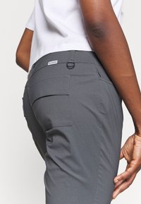 Zbliżenie osoby noszącej szare spodnie turystyczne z tylną kieszenią i małym logo Columbii na pasku.