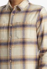 Camisa de franela a cuadros en crema, amarillo y púrpura. Presenta cierre de botones, dos bolsillos en el pecho y un collar clásico. Textura suave.