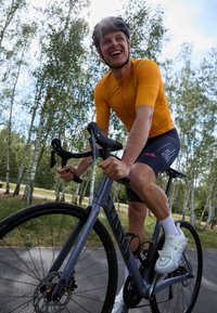 Cycliste sur un vélo de route gris avec freins à disque, portant un maillot à manches courtes orange et un short noir, des chaussures de cyclisme blanches, sur un chemin entouré de verdure.