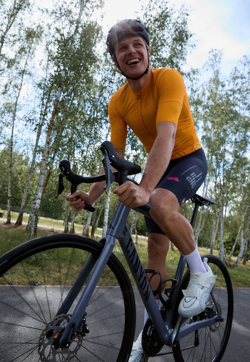 Cycliste sur un vélo de route gris avec freins à disque, portant un maillot à manches courtes orange et un short noir, des chaussures de cyclisme blanches, sur un chemin entouré de verdure.