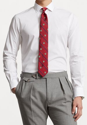 Mann trägt ein weißes Hemd, eine rote gemusterte Krawatte und graue, hochgeschnittene, plissierte Hose mit seitlichen Verstelllaschen.