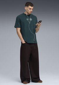 Camiseta de algodón verde oscuro con dos logotipos circulares, combinada con pantalones negros holgados. El personaje sostiene un teléfono inteligente con auriculares.