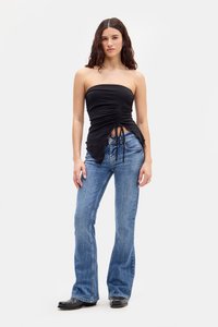 Žena s dlhými kučeravými vlasmi, oblečená v čiernom topu bez ramienok s riasením, modrých rozšírených džínsoch, čiernych topánkach a strieborných prsteňoch, stojí na bielom pozadí.