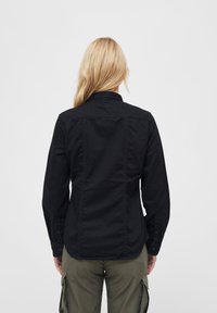 Camicia nera a maniche lunghe con colletto a bottoni, caratterizzata da una texture liscia e un taglio dritto, indossata sopra pantaloni cargo verdi.