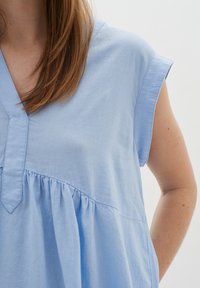 Šviesiai mėlynas, trumpomis rankovėmis viršus su V formos iškirpte ir susiraukšlėtu audiniu liemenyje, pagamintas iš minkštos, tekstūruotos medžiagos.