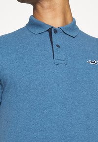 Modrá polo košile s texturovanou tkaninou, s knoflíkovou lištou na tři knoflíky a kontrastním límcem. Na hrudi se nachází malé logo.