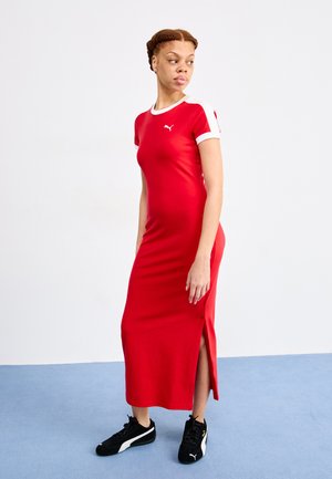 Red jersey maxi dress with short sleeves, white accents, and side slit. Worn with black sneakers, on a blue carpeted surface.