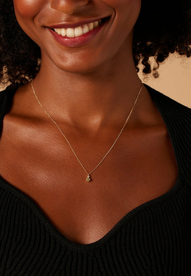 Gouden ketting met een klein groen accent, gedragen door een persoon met een donkere huid, boven een geribd zwart topje met een V-hals.