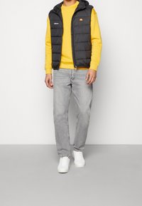 Svart pufferväst med huva över en gul sweatshirt, kombinerad med ljusgrå jeans och vita sneakers. Västet har en logotyp på bröstet.