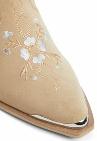 Botín de ante beige hasta el tobillo con bordado floral en hilo plateado, punta afilada y un acento metálico en la puntera. Textura suave.