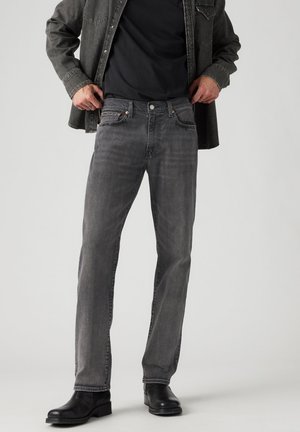 Homme ajustant un jean gris porté avec une chemise noire, une veste gris foncé et des bottines noires sur un fond clair uni.