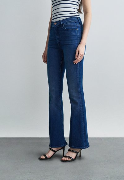 Femme portant un jean bootcut bleu marine, des sandales à talons noires, et un haut sans manches blanc à rayures bleu marine, debout sur un sol gris.