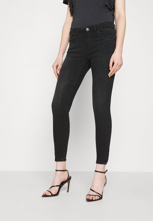 Mujer con jeans ajustados de cintura alta en negro y sandalias negras de tacón con tiras, sobre un fondo blanco liso.