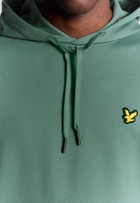 Felpa verde realizzata in tessuto morbido, con cordini con punte nere e un piccolo logo di un uccellino giallo sul petto.