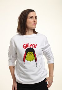 Felpa bianca con un cartone animato del Grinch che indossa un cappello nero e dei paraorecchie rossi. Un testo in grassetto recita "Il GRINCH" sopra il grafico.