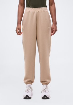 Άτομο που φοράει μπεζ ελαστικό παντελόνι με λάστιχο στη μέση και λευκά αθλητικά παπούτσια, στέκεται μπροστά σε απλό φωτεινό φόντο.