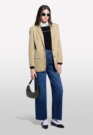 Blazer en daim beige, pull noir, chemise rayée, jeans bleu à jambes larges, sac à main noir avec œillets et chaussures noires à bout pointu avec des chaussettes blanches.