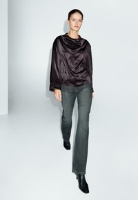Donna che cammina in avanti, indossando un top lungo e lucido di colore scuro, jeans grigi a gamba dritta e stivali neri con tacco, su uno sfondo bianco semplice.