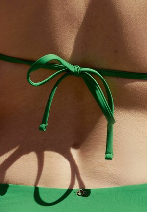 Groen bikini top met een dunne strik, gemaakt van gladde stof en voorzien van een klein gouden embleem aan de onderzijde, tegen een gestructureerde huid achtergrond.