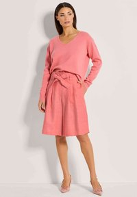 Rosa V-Ausschnitt-Pullover und hochgeschnallte rosa Shorts mit einem Bindedetail. Das Outfit zeichnet sich durch eine weiche Textur und eine entspannte Passform aus.