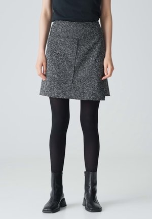 Femme portant une mini-jupe grise texturée, des collants noirs, des bottines noires et un haut noir, debout devant un fond clair uni.