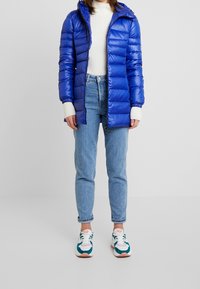 Chaqueta azul acolchada con capucha, puños acanalados y cierre de cremallera, combinada con jeans de color azul claro y zapatillas multicolor.