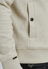 Sweatshirt en polaire beige avec un motif alvéolé texturé, détail de bouton sur la poche, poignets et ourlet côtelés, et petite étiquette de marque sur la manche.