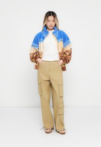 Une femme se tient avec les mains dans les poches, portant une veste en polaire multicolore, une chemise blanche, un pantalon cargo beige et des baskets beiges sur fond blanc.