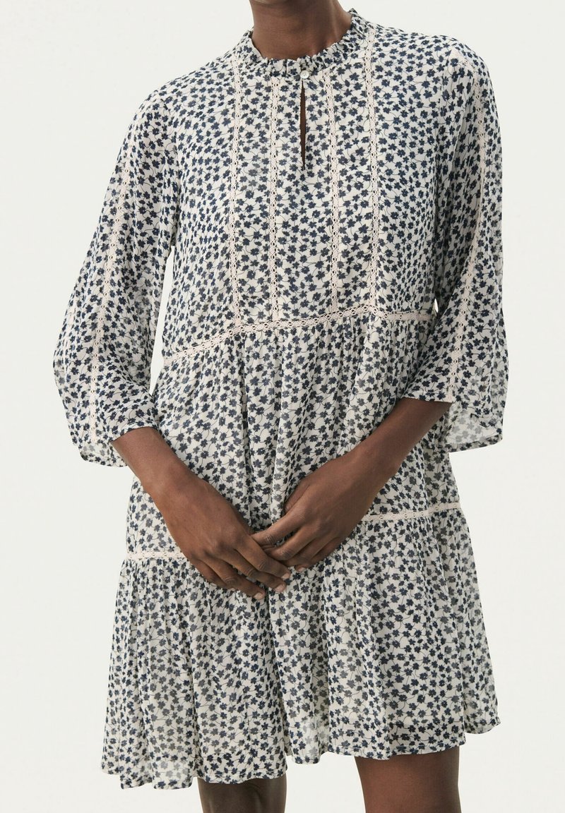 Personne portant une robe blanche longueur genou avec de petits motifs floraux noirs et des détails en dentelle, debout, les mains doucement jointes devant.