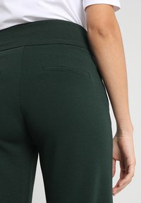 Mörkgröna, högmidjade byxor med en slät yta och en figurnära design. Har två små bakfickor och en ren sömnadsavslutning.