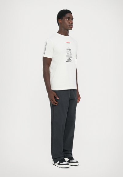 Camiseta de algodón blanca con estampados gráficos en rojo y negro, mangas cortas y un corte relajado, combinada con pantalones gris oscuro y zapatillas negras.